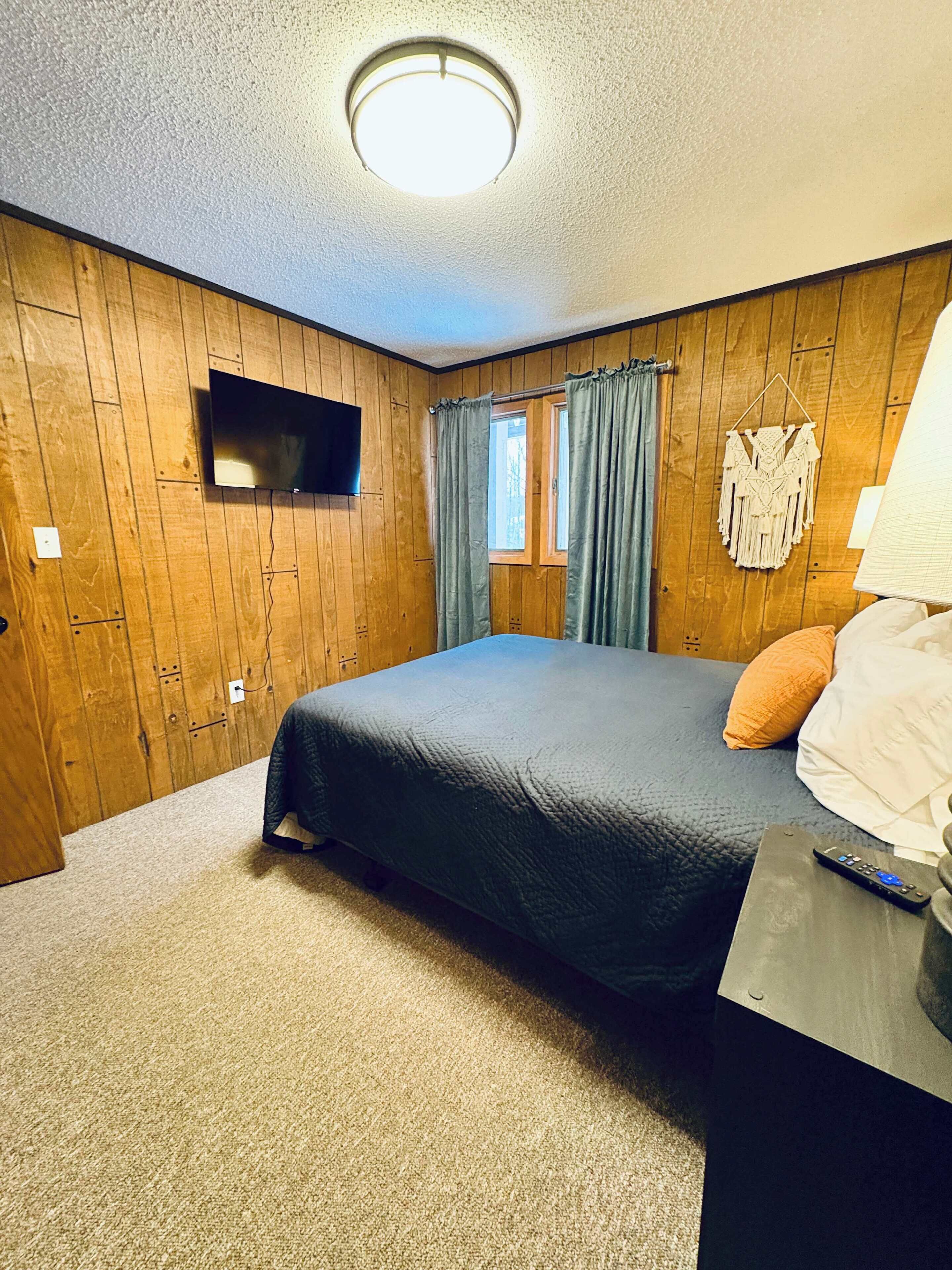 A bedroom with a bed, a TV mounted on the wall, a window with curtains, and a lamp on a bedside table.