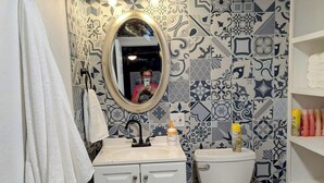 A bathroom with a white sink, a toilet, and a mirror. There are patterned tiles on the walls and a shelf with toiletries. - The Half Pint - Pet Friendly for 1 Pet Only (Abilene)