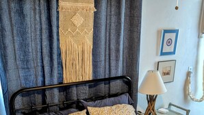 A bed with a patterned bedspread and pillows, a bedside lamp, a small table with a lamp, and a macramé wall hanging. - The Half Pint - Pet Friendly for 1 Pet Only (Abilene)