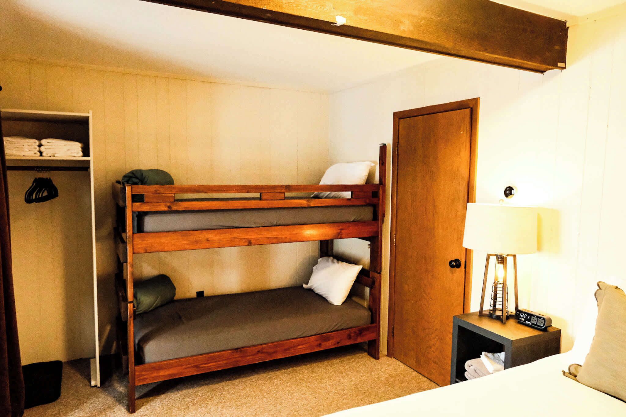 A bunk bed with a wooden frame, a bedside table with a lamp, and a wardrobe with folded clothes.