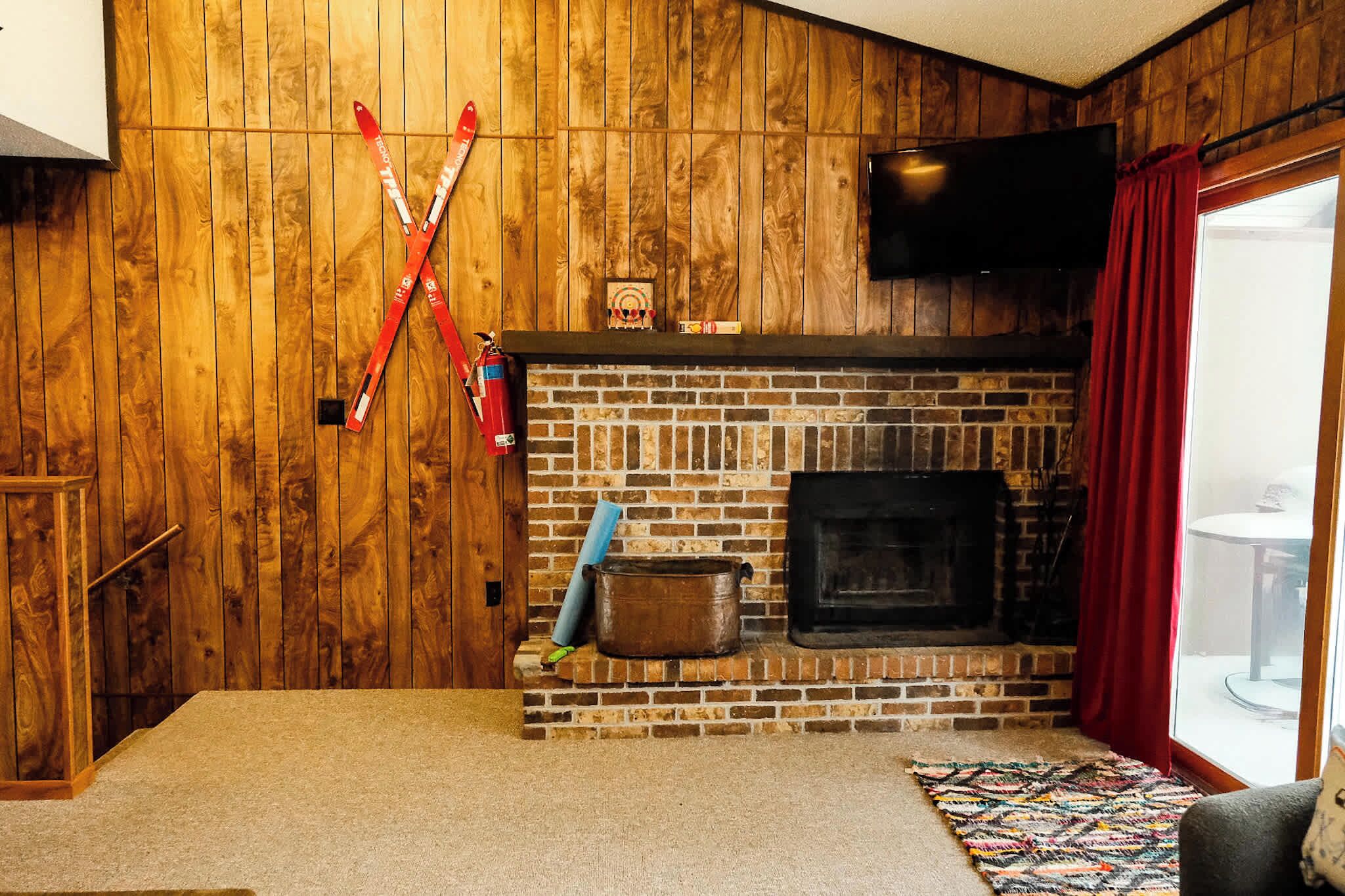 A room with a brick fireplace, wooden walls, a television mounted above it, and a red curtain on the window.