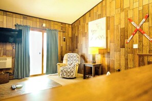 A living room with wooden paneling, a television, a chair, a table with a lamp, and a large painting.