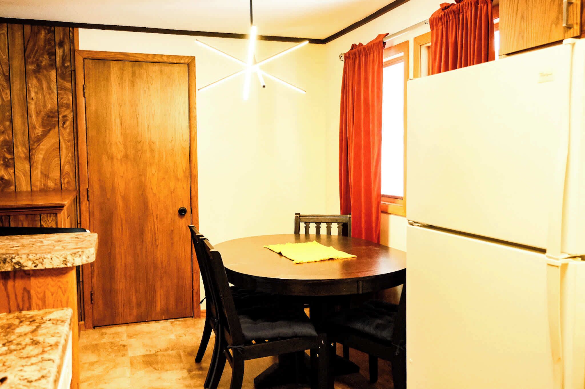Una cucina compatta con una porta in legno, un tavolo da pranzo con sedie, un frigorifero e una lampada a sospensione moderna.