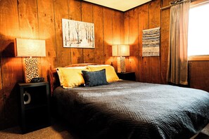 Une chambre à coucher aux murs lambrés, avec un lit, deux tables de chevet dotées de lampes, une œuvre d’art accrochée au mur et une fenêtre avec des rideaux.