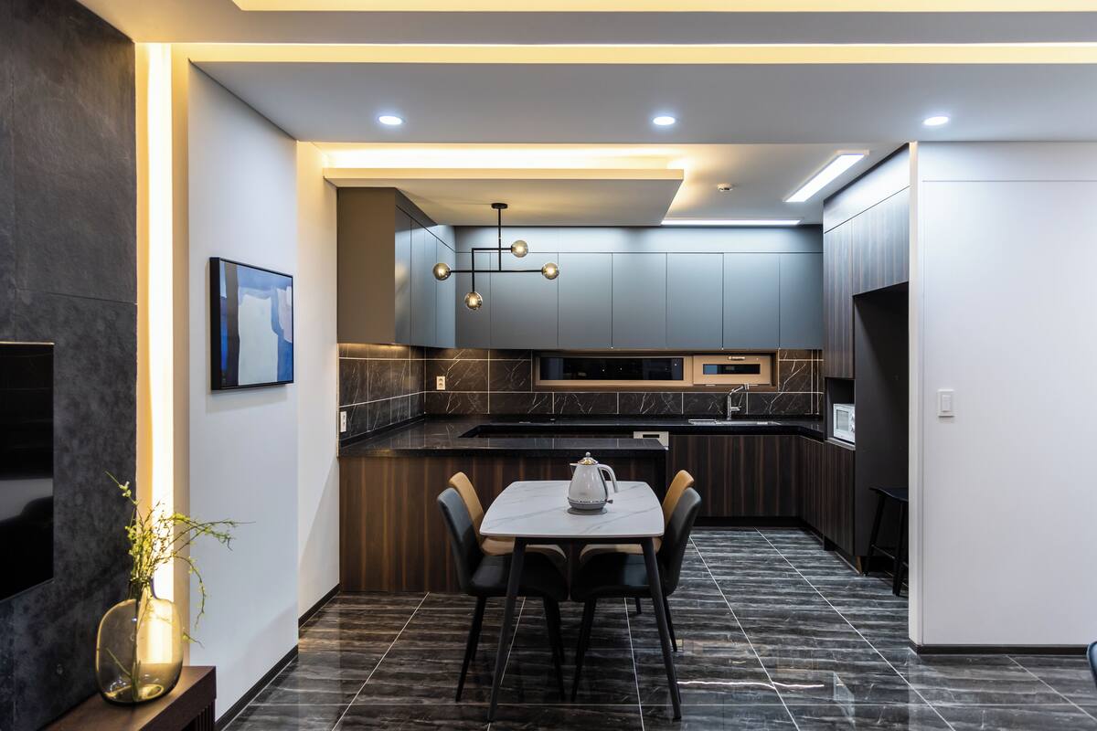 A modern kitchen with dark cabinetry, a marble countertop, and a dining area with a table and chairs.