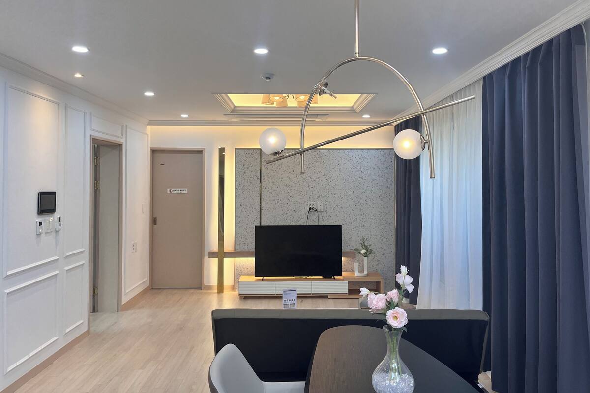 A modern living room with a flat-screen TV, a coffee table with a vase of flowers, and a dining area with a chair.