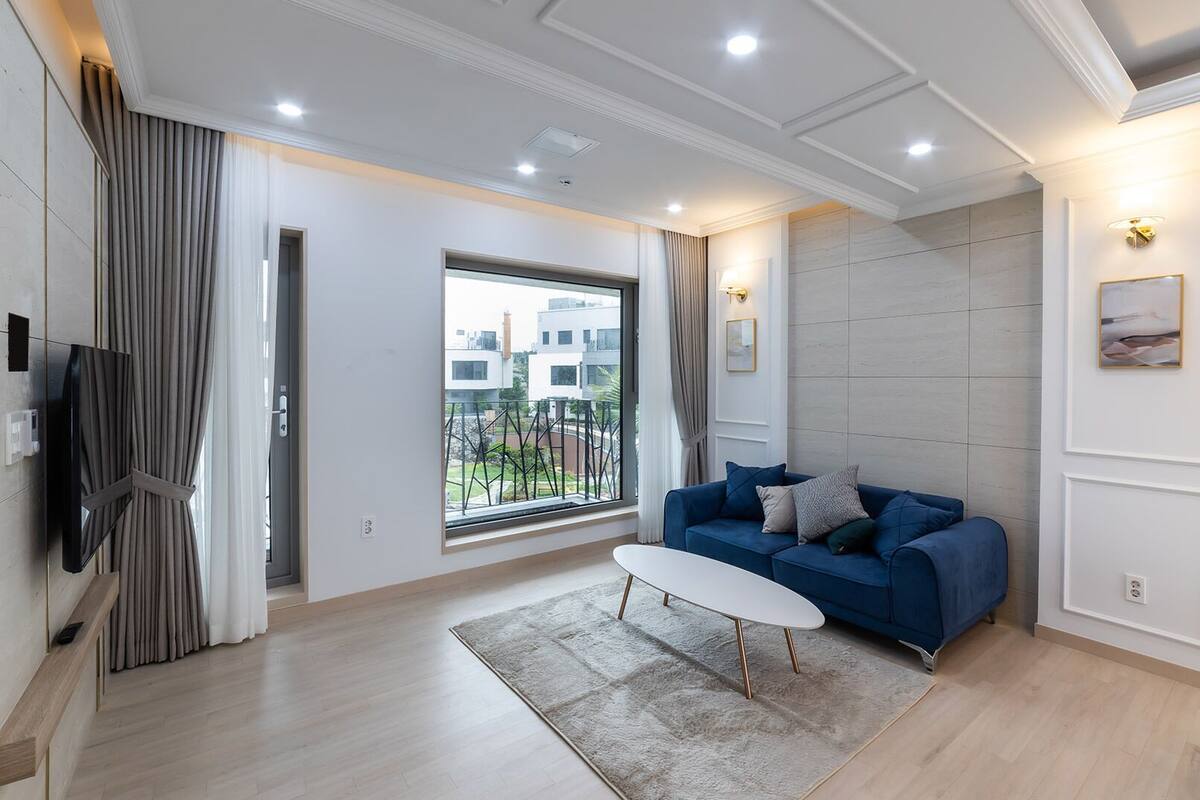 A modern living room with a blue sofa, a white coffee table, a large window with a view, and recessed ceiling lighting.
