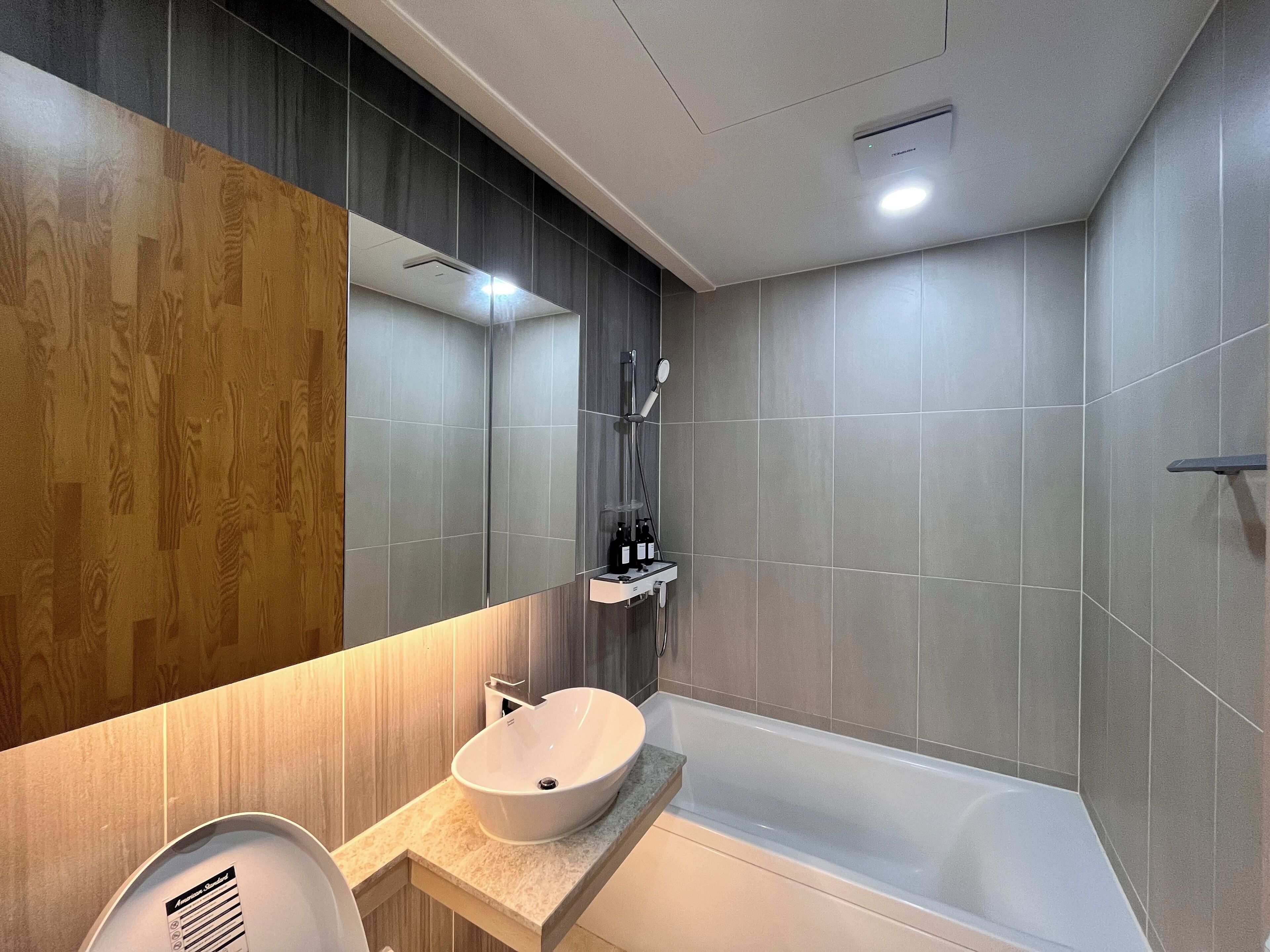 A modern bathroom with a wooden vanity, a large mirror, a bathtub, and grey tiles.