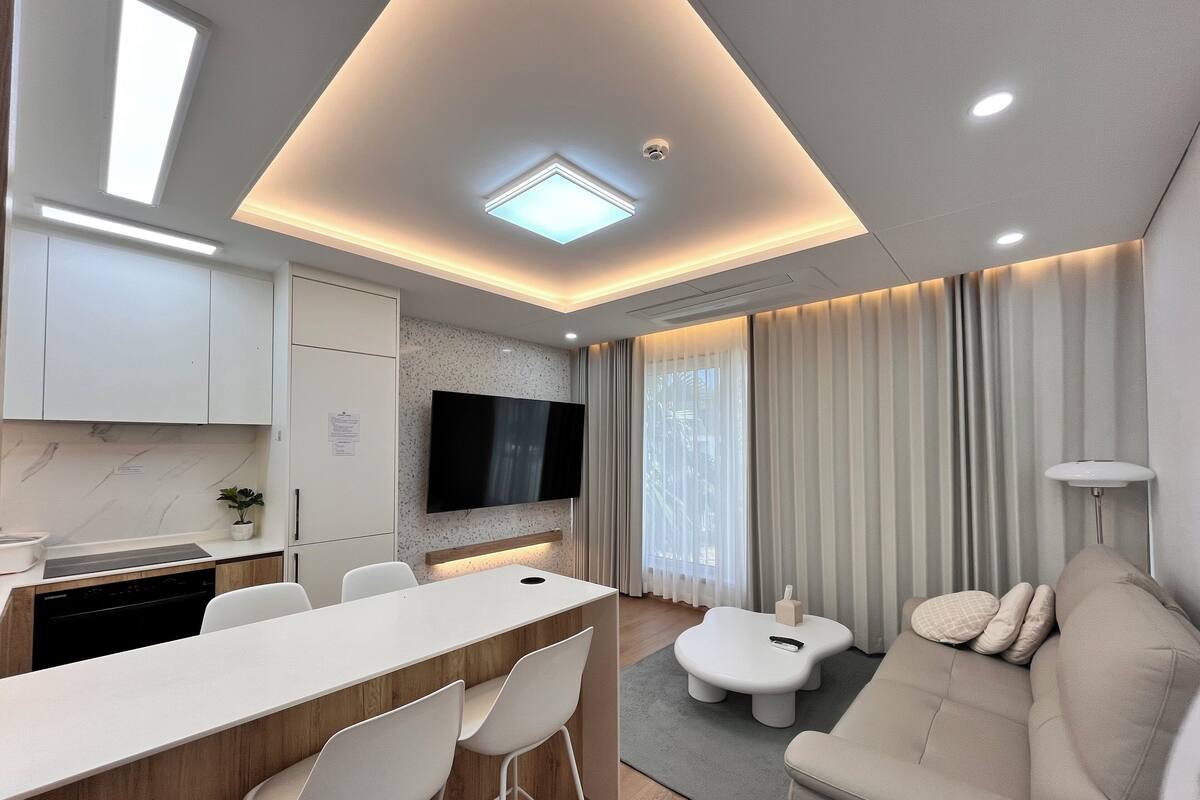 A modern living room with a sofa, a dining area with chairs, a TV mounted on the wall, and a kitchen area with cabinets and a countertop.