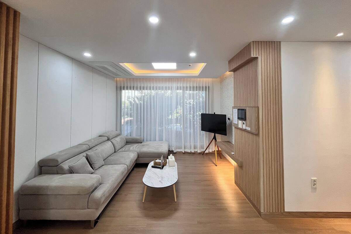 A modern living room with a sectional sofa, a coffee table, a flat-screen TV on a stand, and a large window with sheer curtains.