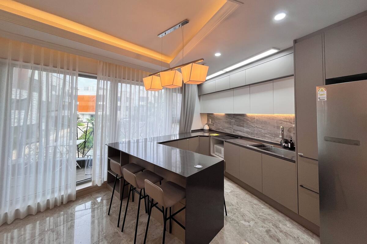 A modern kitchen with a central island, bar stools, stainless steel appliances, and a view of the outdoors through large windows.