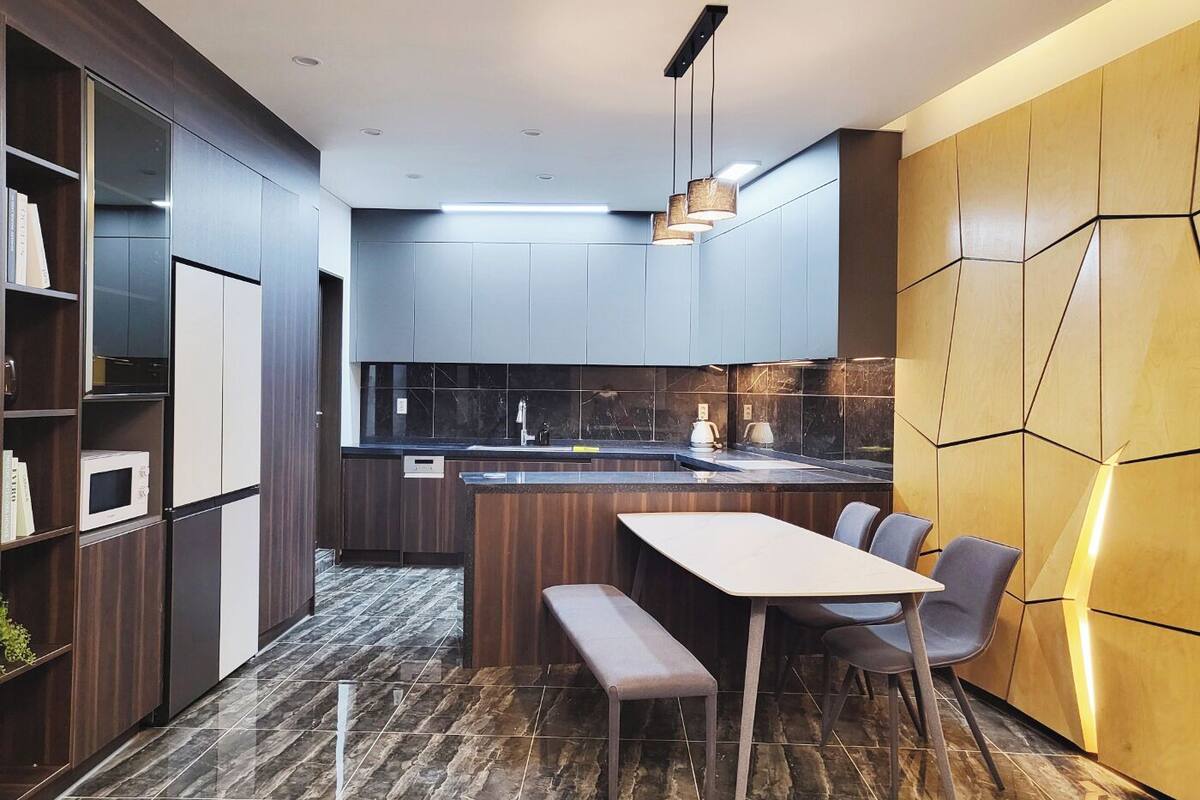 A modern kitchen with dark cabinetry, a marble countertop, and a dining area with a bench and chairs.