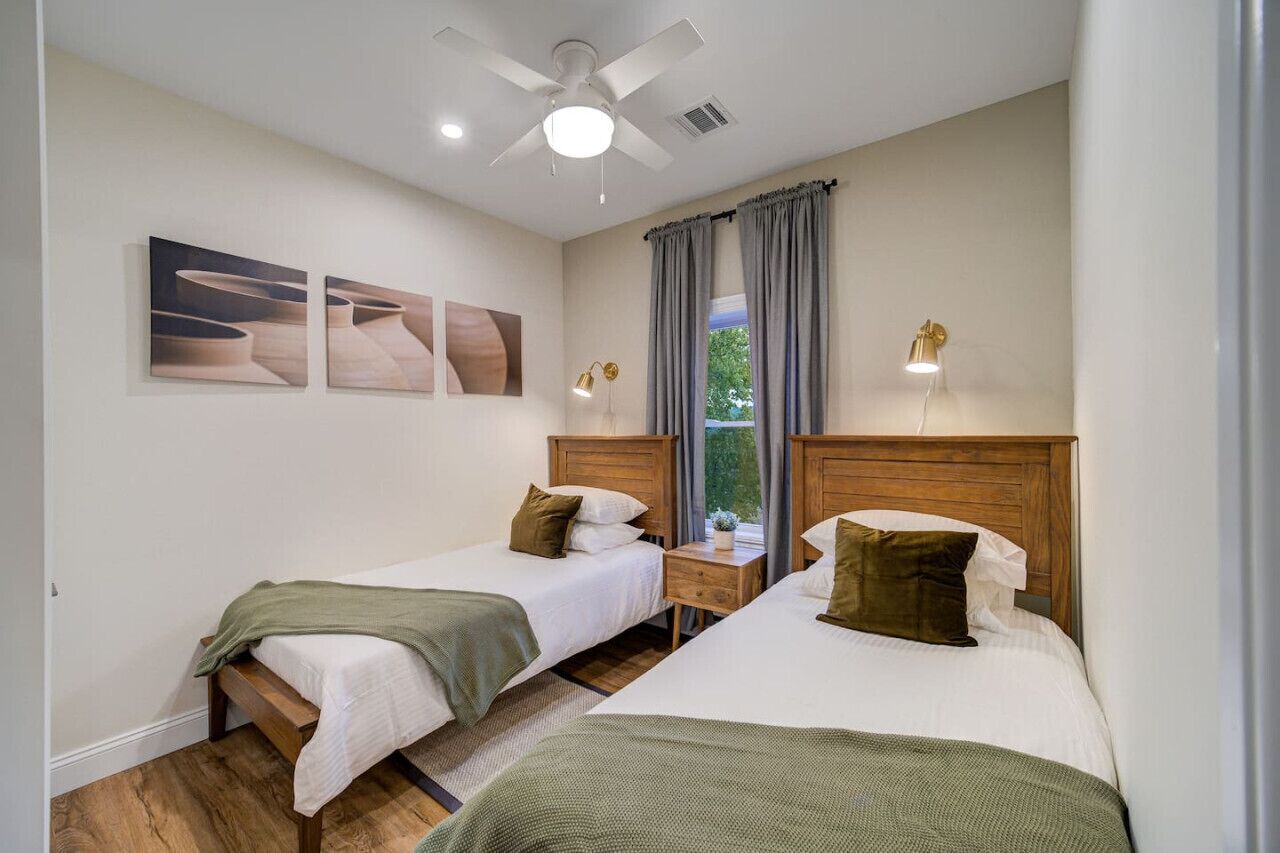 Une chambre d’hôtel avec deux lits simples, un ventilateur de plafond, une fenêtre avec des rideaux et des œuvres d’art accrochées au mur.