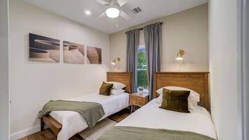 Une chambre d’hôtel avec deux lits simples, un ventilateur de plafond, une fenêtre avec des rideaux et des œuvres d’art accrochées au mur.