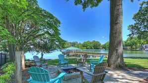 An outdoor seating area with Adirondack chairs and a fire pit, overlooking a lake and dock. - Lakeshore Lookout - Amazing Long Range Views, Pool, Hot Tub, Fire Pit, Theatre Room and much more! (Mooresville)