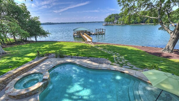 A swimming pool with a hot tub, surrounded by a stone deck and a grassy area with a dock extending into a lake. - Lakeshore Lookout - Amazing Long Range Views, Pool, Hot Tub, Fire Pit, Theatre Room and much more! (Mooresville)