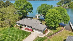 A large house with a garage, surrounded by a well-maintained lawn and trees, near a lake. - Lakeshore Lookout - Amazing Long Range Views, Pool, Hot Tub, Fire Pit, Theatre Room and much more! (Mooresville)