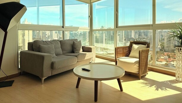 Una sala de estar moderna con sofá, sillón y mesa de centro, ventanas amplias con vista a la ciudad.