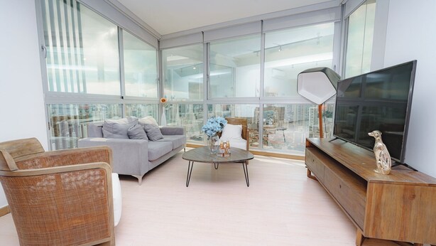 A modern living room with a grey sofa, wicker chair, and a coffee table with flowers. A TV is on a wooden stand, and large windows offer a city view.