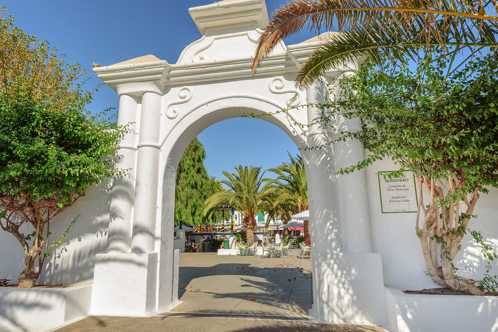 An ornate white archway with a plaque, surrounded by greenery and palm trees.