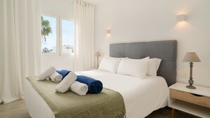 A neatly made bed with a green throw, white bedding, and rolled towels. There is a nightstand with a lamp and a wall-mounted light. A window with white curtains allows natural light in. - Lanzahost Tabaiba Green (Costa Teguise)
