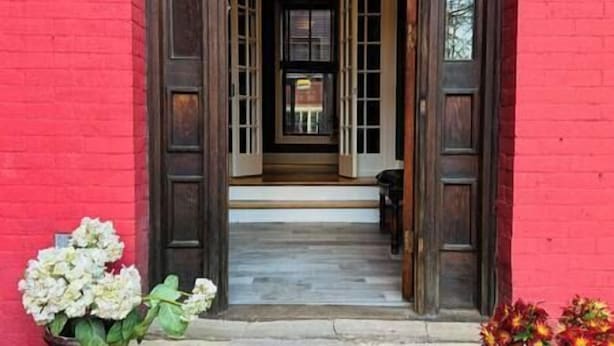Un edificio in mattoni rossi con una porta d'ingresso in legno, un piccolo portico e un vaso con fiori bianchi.