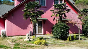 Une maison rose avec un toit en pente, deux fenêtres et une porte. On y trouve deux palmiers et un arbuste dans le jardin.