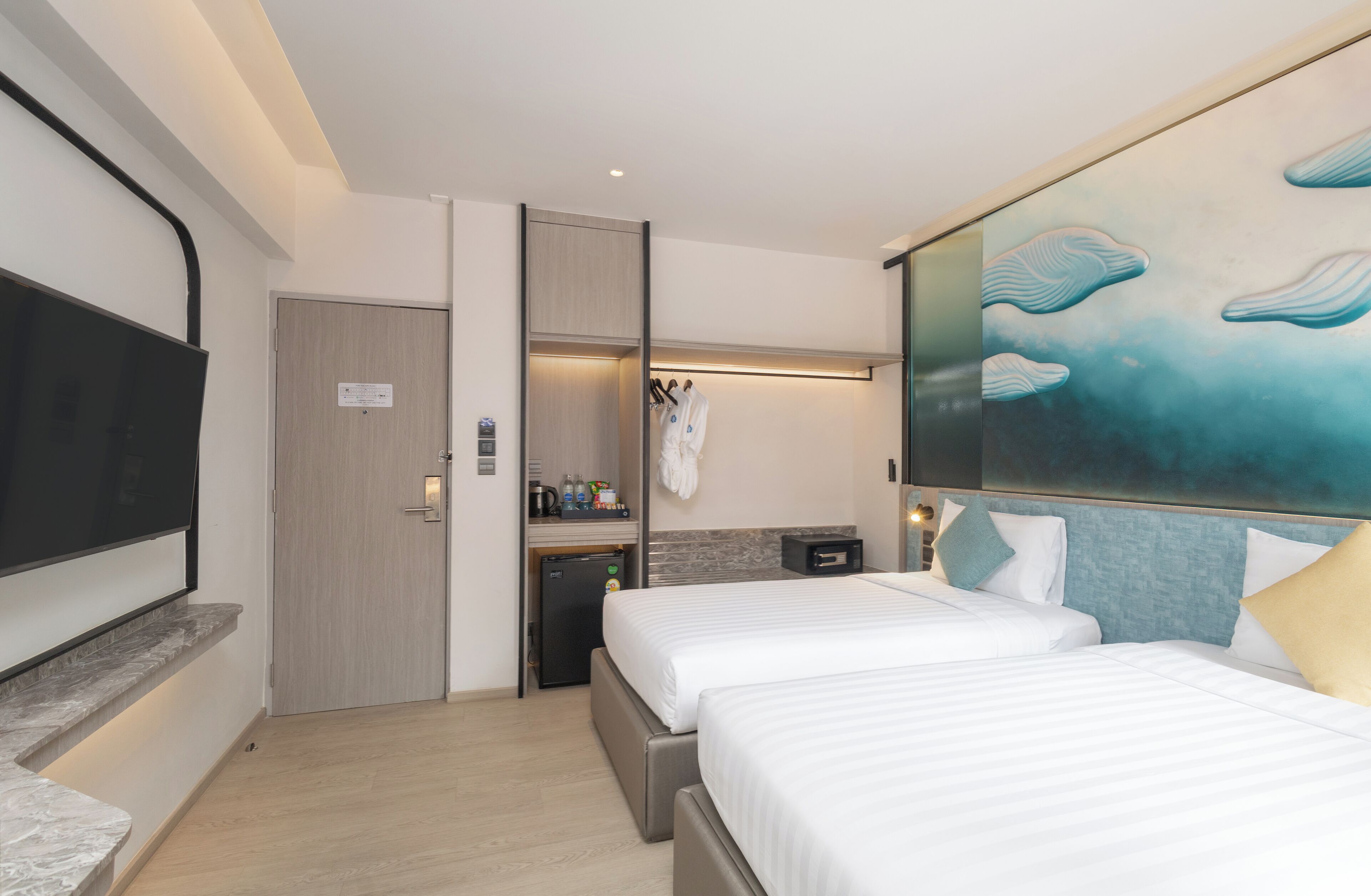 A hotel room with two beds, a flat-screen TV, a small kitchenette, and a bathroom with a towel rack.