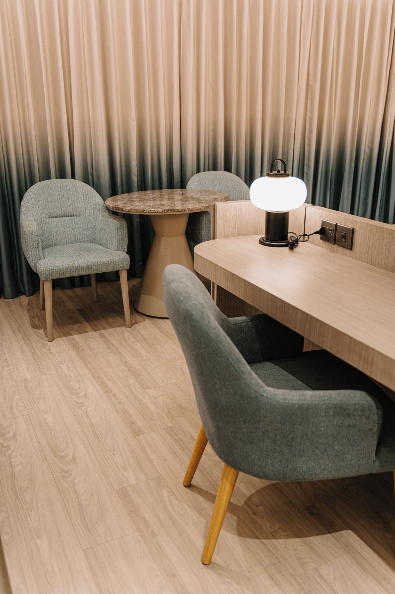 Una habitación con un escritorio de madera, dos sillas grises, una mesa redonda y una lámpara.