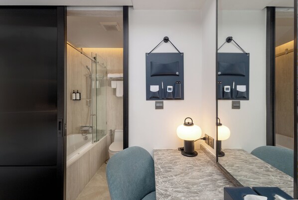 A modern bathroom with a glass shower enclosure, a bathtub, and a toilet. There is a black refrigerator, a marble countertop, and a table with two chairs. Two wall-mounted lamps provide lighting. - Cloudotel Walking Street Pattaya (Pattaya)