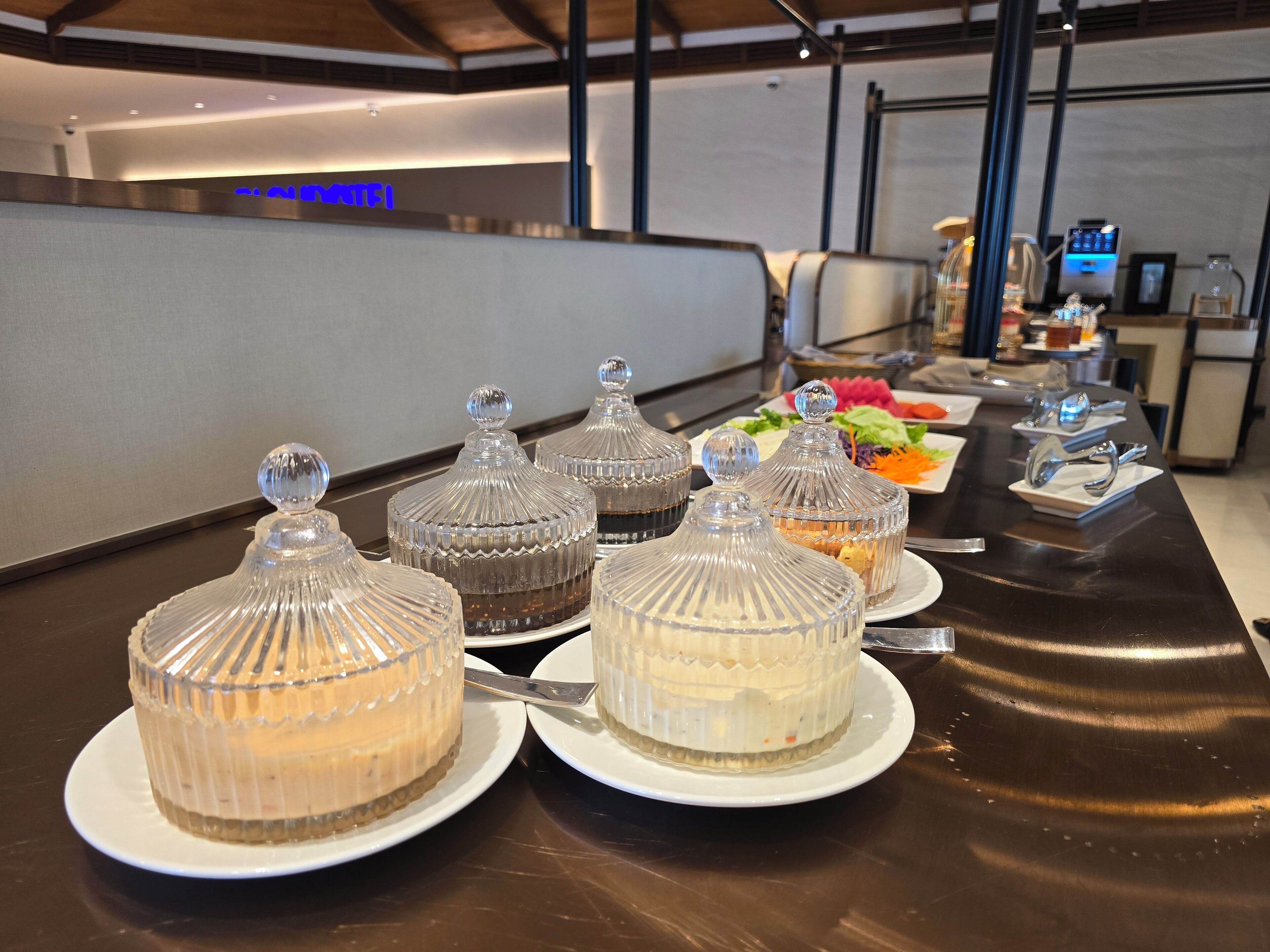 A buffet table with various desserts in glass containers, a salad bar, and a coffee machine in the background.