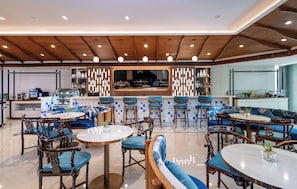Un restaurante moderno con vigas de madera en el techo, mesas redondas, sillas con cojines azules y una zona de bar con un espejo grande.