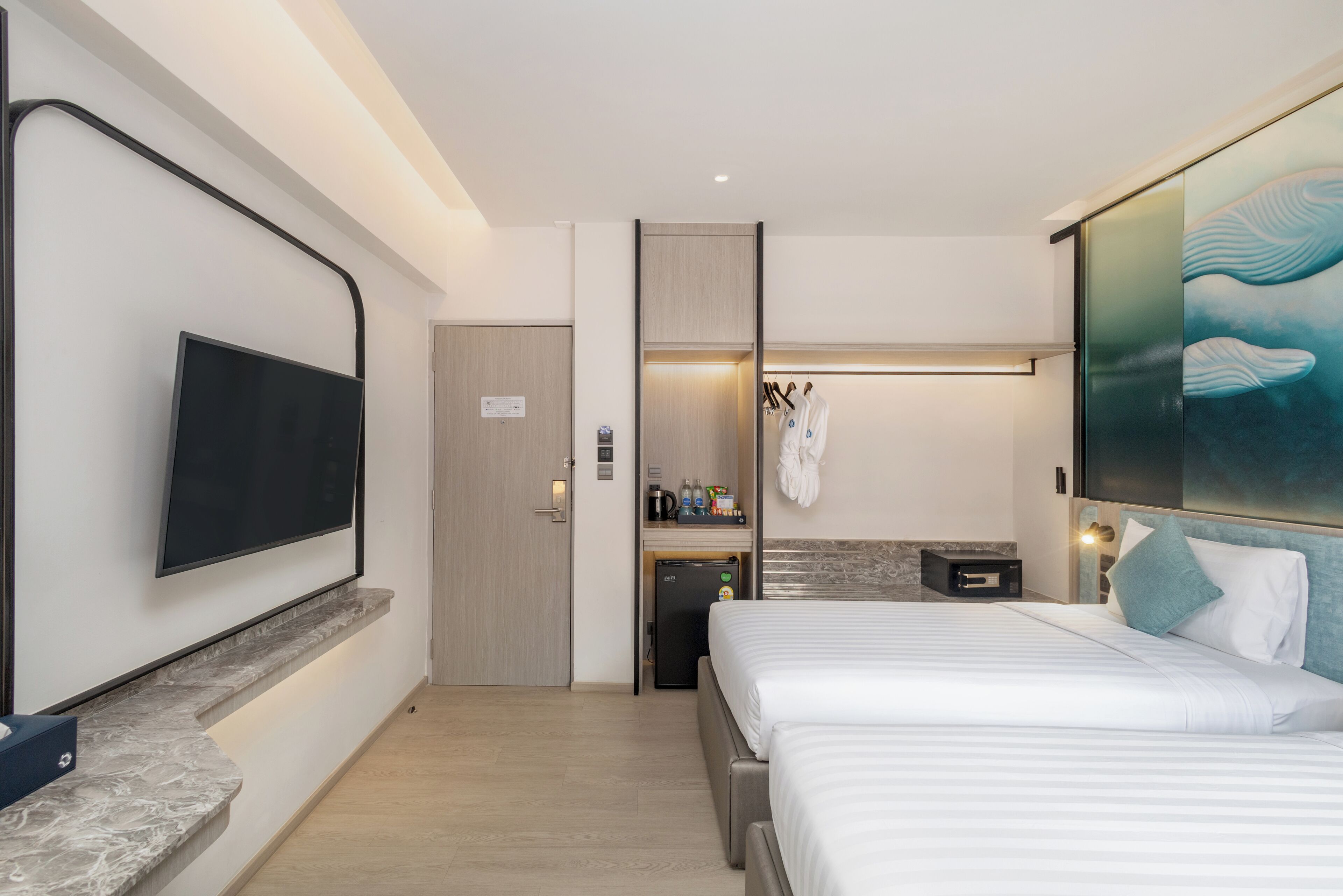 Une chambre d’hôtel moderne équipée d’un téléviseur à écran plat fixé au mur, d’un lit avec une literie blanche, d’un petit bureau avec un ordinateur et d’un mini-bar.