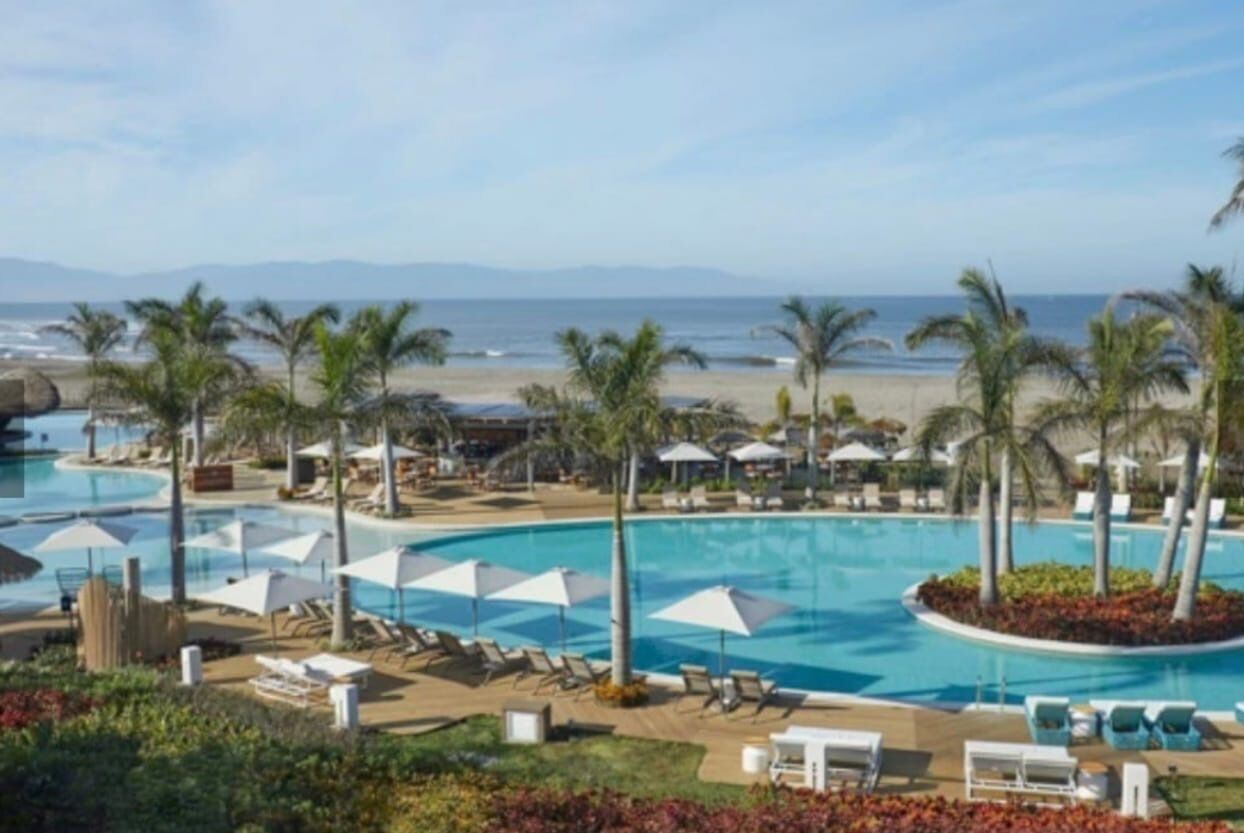 A resort pool area with lounge chairs and umbrellas, a beach view, and palm trees.