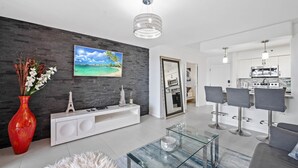 Un salon moderne avec un canapé blanc, une table basse en verre, un vase rouge et un tableau représentant une scène de plage.