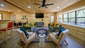 A log cabin interior with a central seating area, a television mounted on the wall, and a large window offering a view of the outdoors. - Whispering Trails Lodge – Secluded Pet-Friendly Broken Bow Cabin | Sleeps 9 | Forest Trails Near Lake & Beavers Bend (Hochatown)