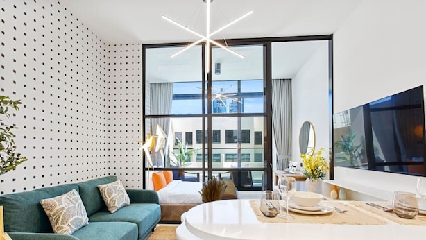 A modern hotel room with a green sofa, a round dining table, and a large window offering a city view. - Sleek Brand New Studio in Loci Residence JVC (Dubai)
