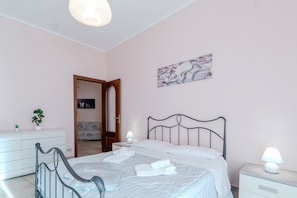 A bedroom with a bed, bedside tables, a nightstand, a wall-mounted lamp, a door leading to another room, and a small potted plant.