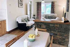 Un salon chaleureux avec une cheminée en pierre, une table à manger ornée d’un panier de fruits et un canapé recouvert d’une couverture.