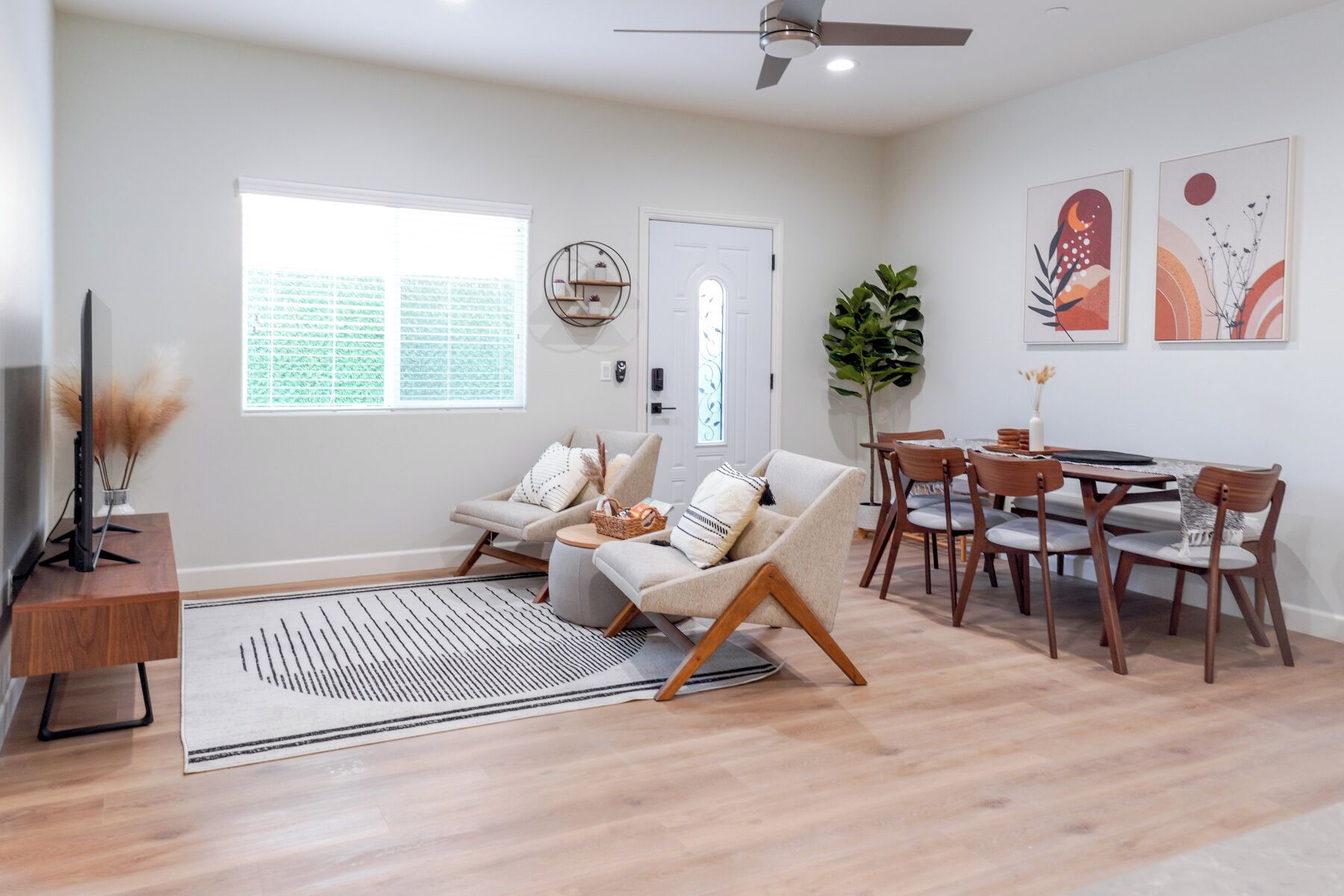 A spacious living area with a sofa, a dining table with chairs, a TV on a stand, and a large window with blinds.