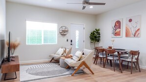 Un vaste espace de vie comprenant un canapé, une table à manger avec des chaises, un téléviseur posé sur un meuble et une grande fenêtre équipée de stores.