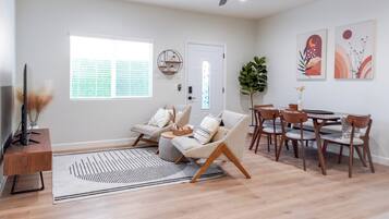 A spacious living area with a sofa, a dining table with chairs, a TV on a stand, and a large window with blinds.