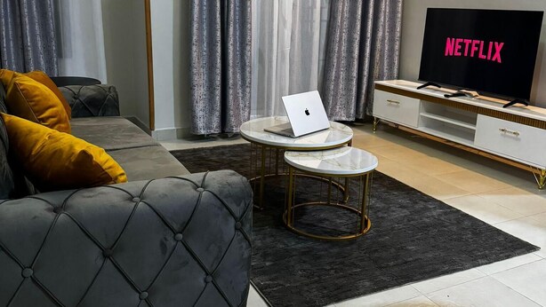 Una sala moderna con un sofá gris, una pequeña mesa redonda de centro, una computadora portátil y un televisor mostrando 'NETFLIX'.