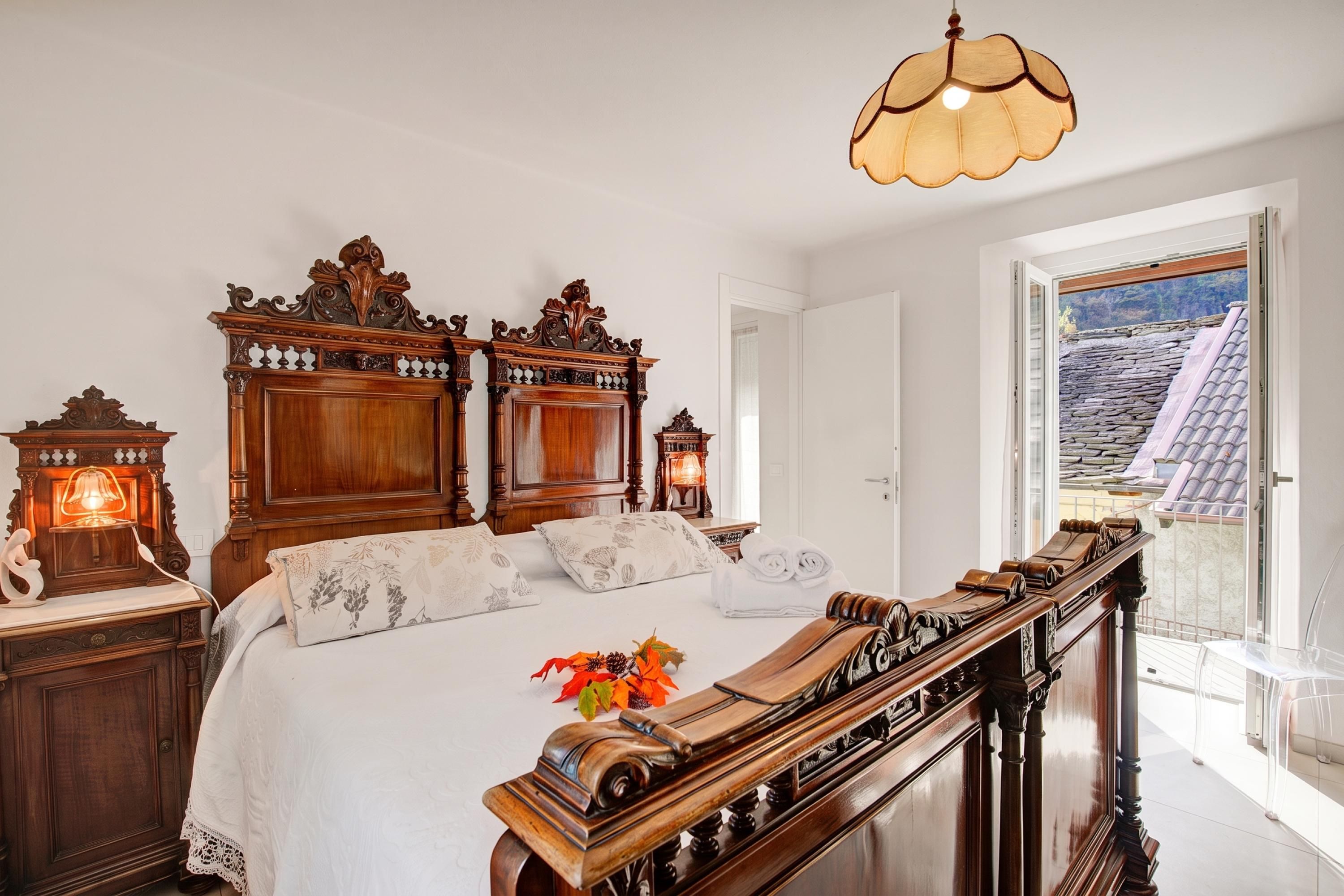 Un dormitorio con una cama grande, un elaborada cabecera de madera, una mesita de noche con una lámpara, y vistas a un tejado de baldosas a través de una puerta abierta.
