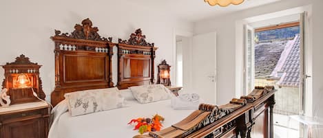 Un dormitorio con una cama grande, un elaborada cabecera de madera, una mesita de noche con una lámpara, y vistas a un tejado de baldosas a través de una puerta abierta.