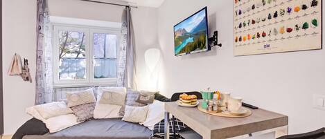 A modern living room with a grey sofa, a flat-screen TV, a dining table with chairs, and a wall-mounted picture of various objects.