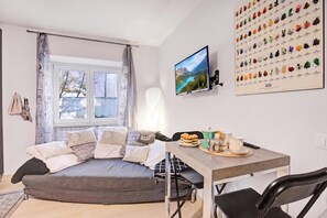 A modern living room with a grey sofa, a flat-screen TV, a dining table with chairs, and a wall-mounted picture of various objects.