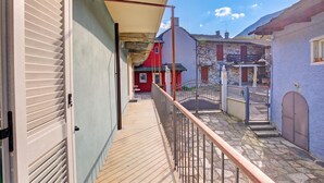 Un balcon étroit doté d’un plancher en bois, d’une rambarde en métal et offrant une vue sur un bâtiment rouge et des maisons en pierre en arrière-plan.
