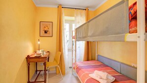 Une petite pièce comprenant un lit superposé, un bureau avec une chaise, un tableau encadré au mur et une fenêtre avec des rideaux.
