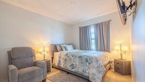 Een slaapkamer met een bed, een grijze fauteuil, twee nachtkastjes met lampen, een raam met gordijnen en een plafondlamp.
